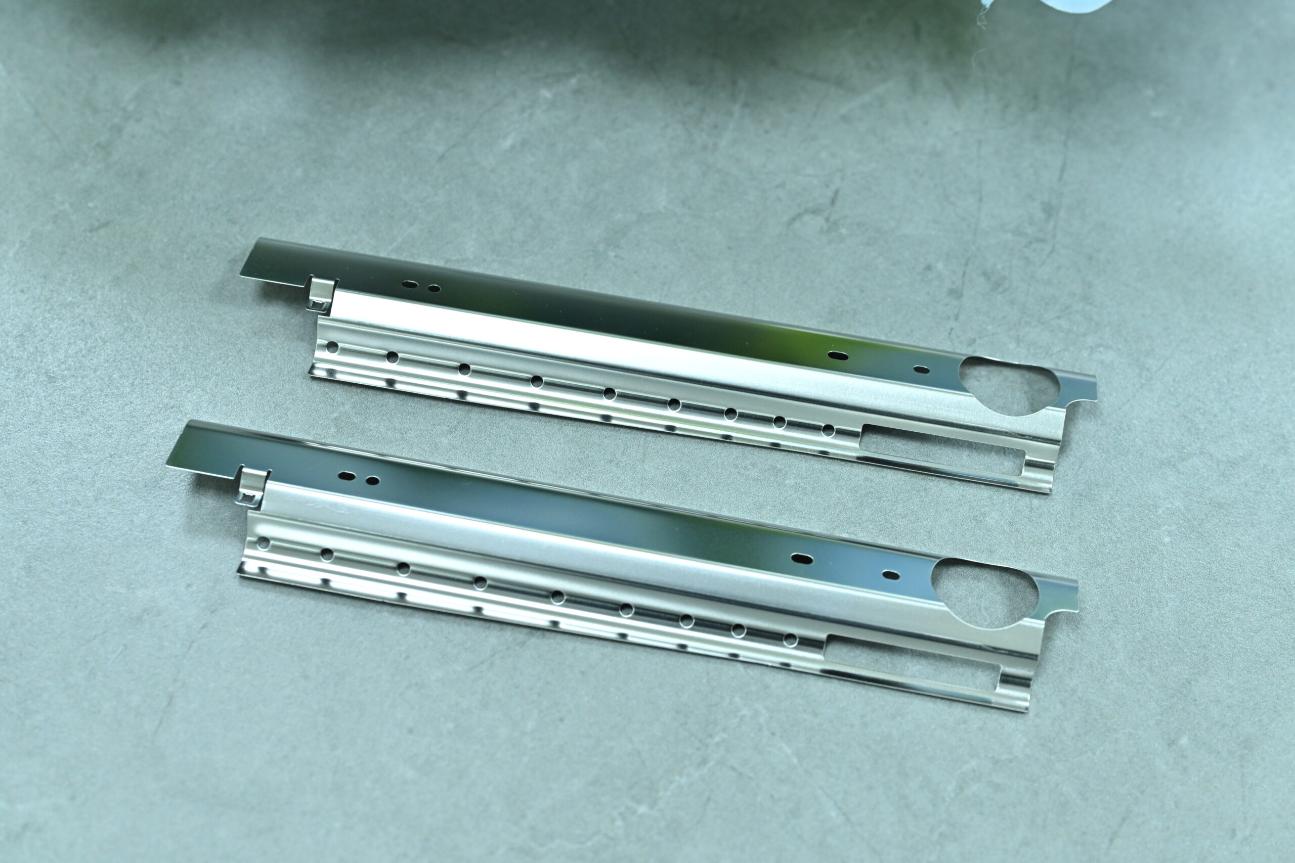 Two metal bracket-like components with holes and slots lie parallel on a smooth gray surface.