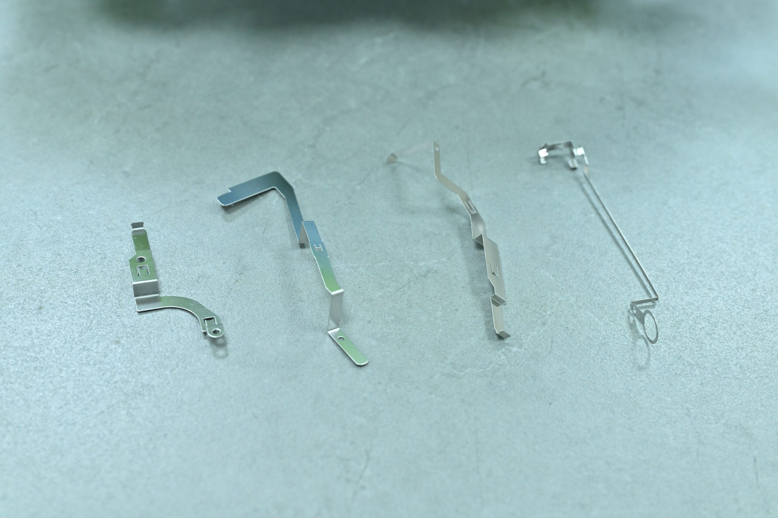 Four thin, silver metal components with varied shapes are arranged in a row on a smooth, light gray surface.