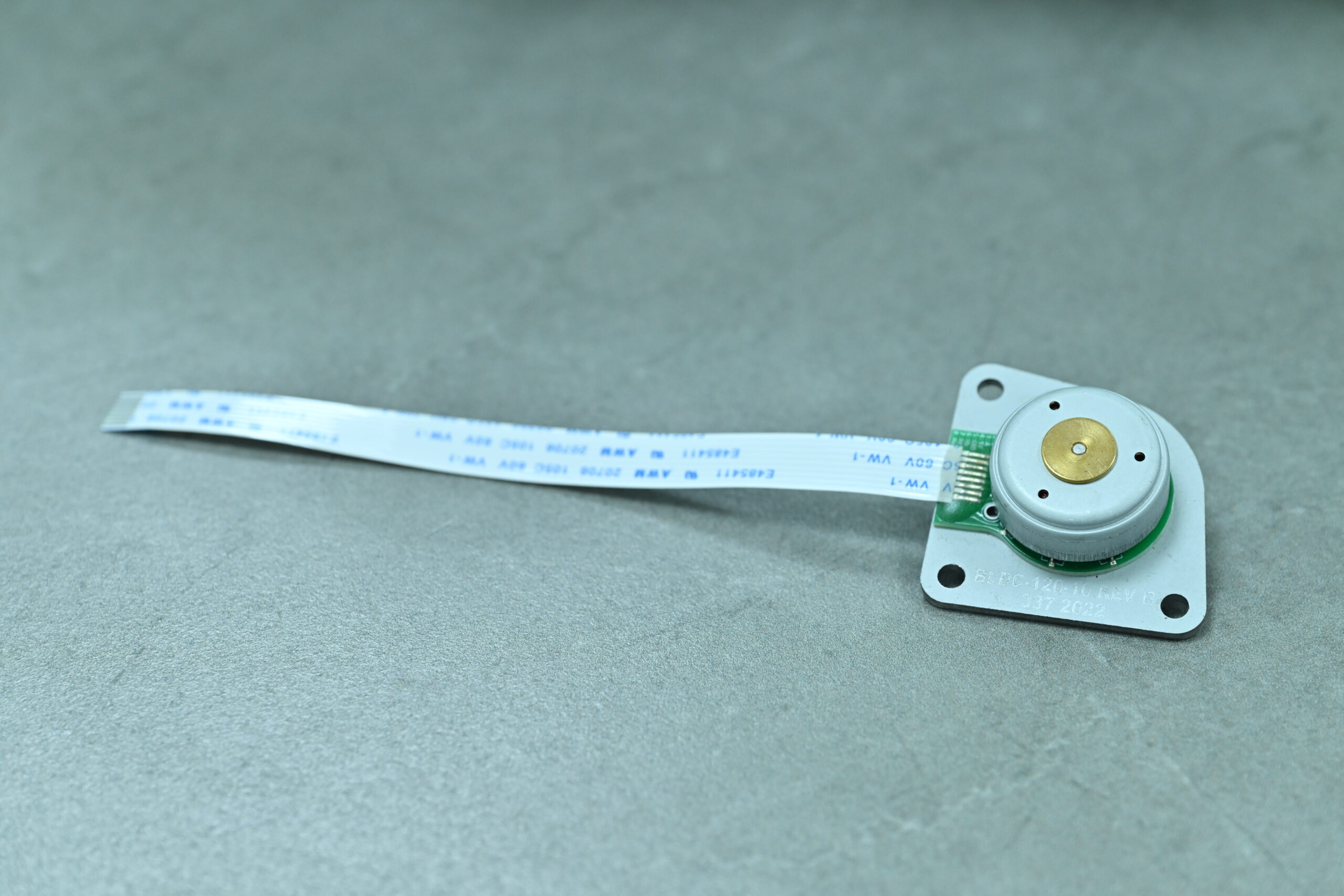 A small electronic component with a ribbon cable is placed on a gray surface. The component has a circular metal part and a white base with four mounting holes.