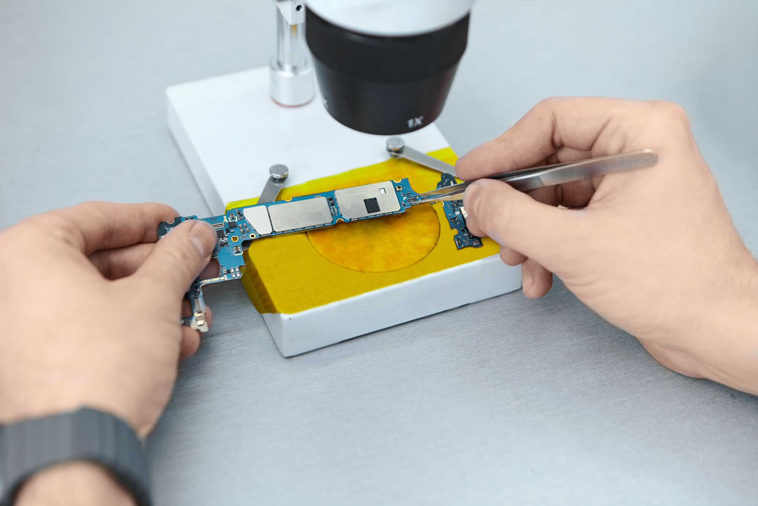 A person uses tweezers to examine a circuit board under a microscope on a workbench.