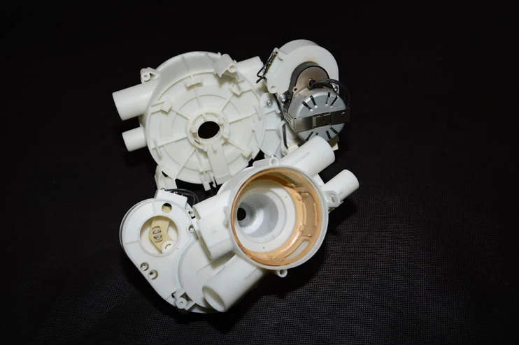 A white plastic mechanical component with cylindrical extensions and a metal part, displayed against a black background.