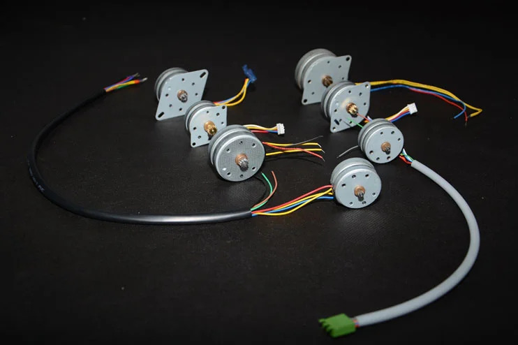 Six small electric motors with attached wires and connectors are arranged on a black surface.