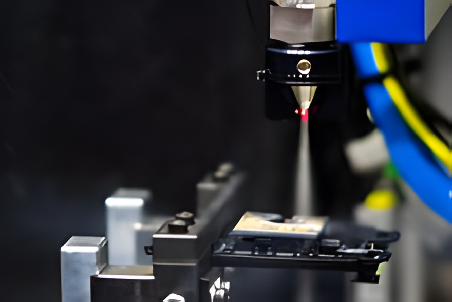 Close-up of an industrial laser machine precisely cutting or engraving a metallic component, with mechanical parts and blue cables visible in the background.