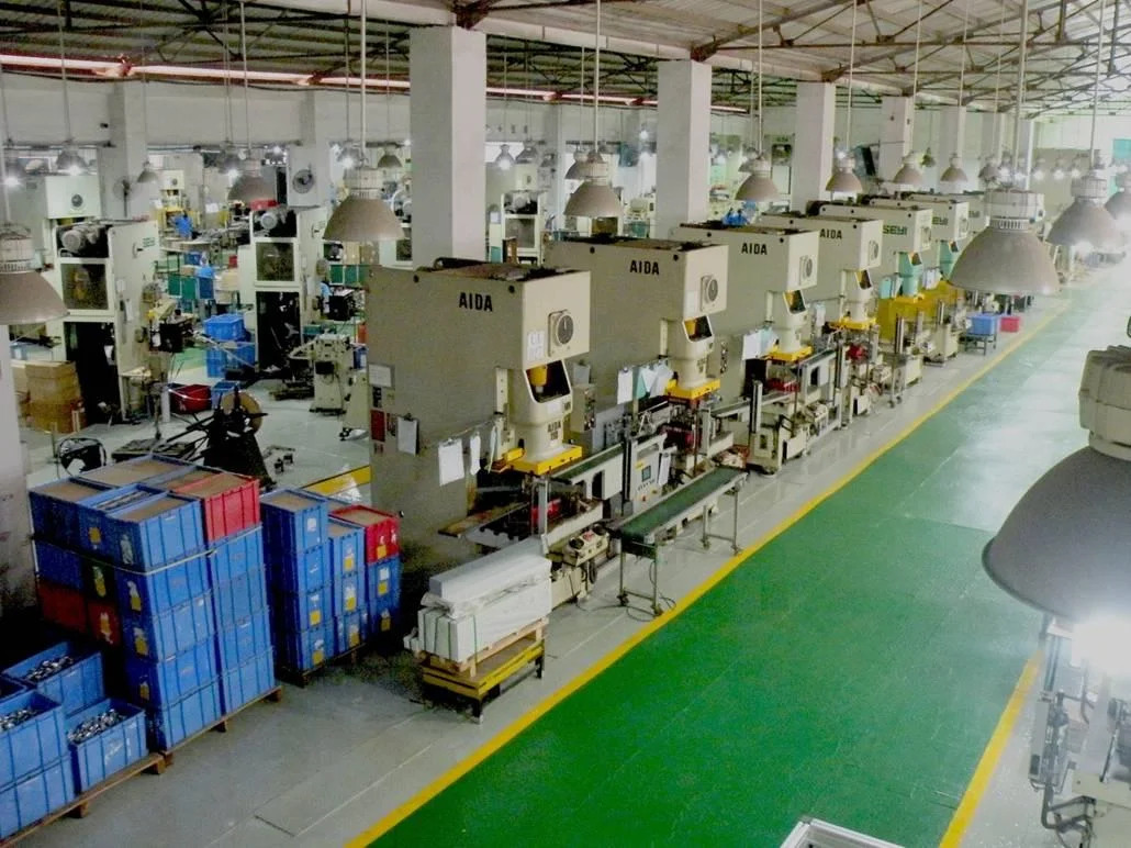 Large industrial factory floor with multiple automated AIDA machines, stacked blue storage bins, and overhead lighting.