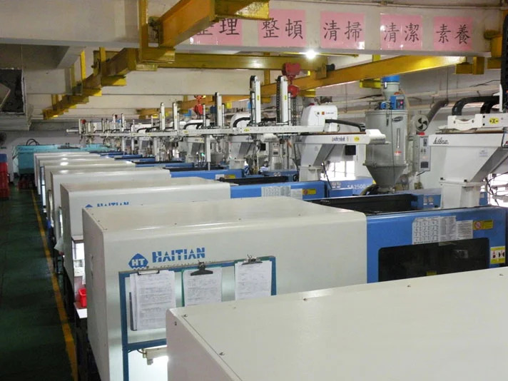 A row of automated injection molding machines operating in a well-lit factory setting, with yellow overhead support beams and control panels visible.