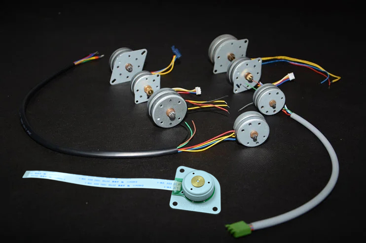Seven small electric stepper motors with attached multicolored wires are arranged on a black background, along with two flat ribbon cables.